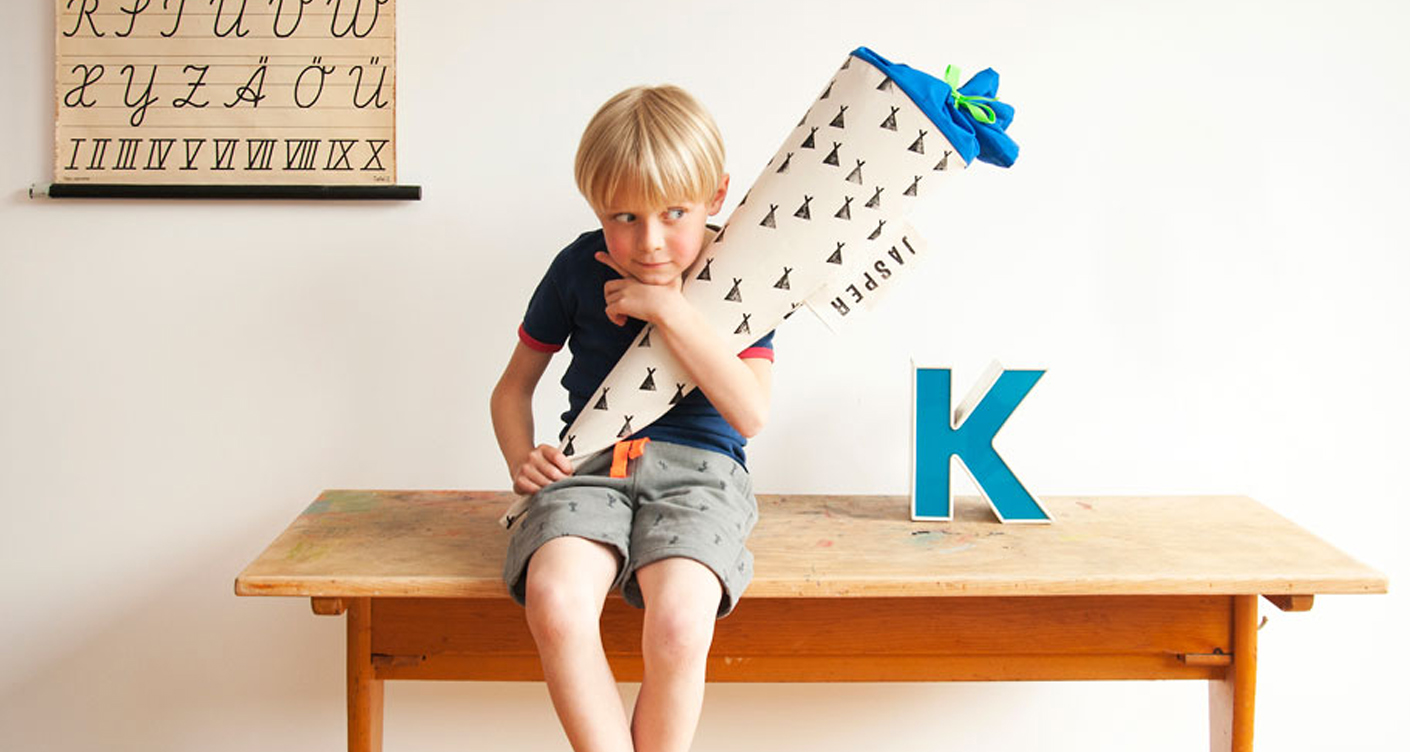 Schultuete von tinyday Kind hält eien Schultuete im Arm und sitzt auf einem Holztisch Buchstaben personalisierbare Zuckertuete Stempel Zelt Einschulung der erste Schultag Schulanfänger
