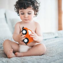 Kleinkind sitzt auf Bett, nur mit Windel bekleidet und spielt mit einem roten Auto